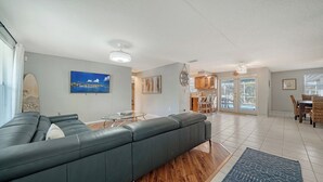 A modern living room with a sectional sofa, a dining area with wicker chairs, and a kitchen visible through an open door.