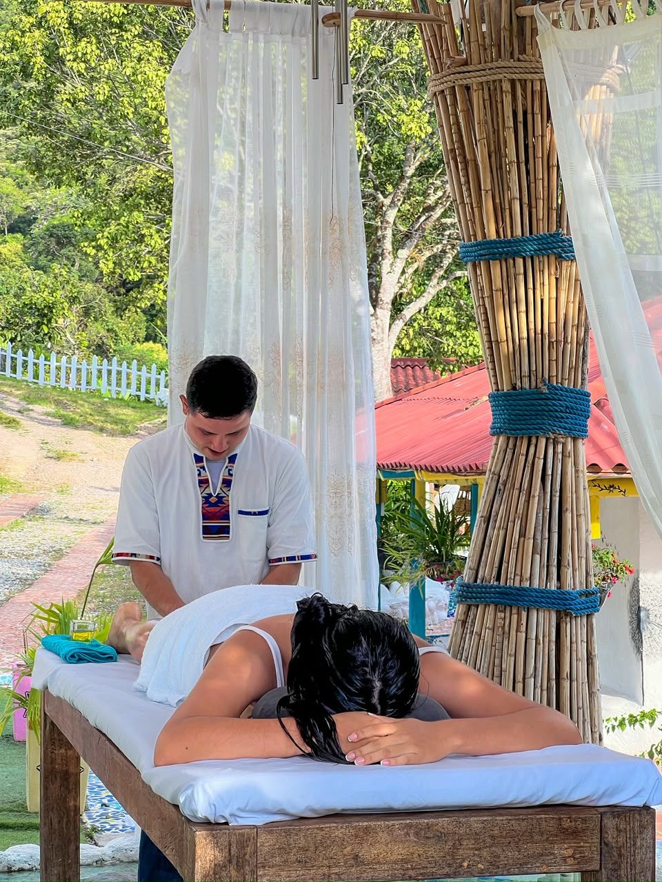 A massage therapy session taking place outdoors with a wooden massage table, a white canopy, and a bamboo structure in the background.