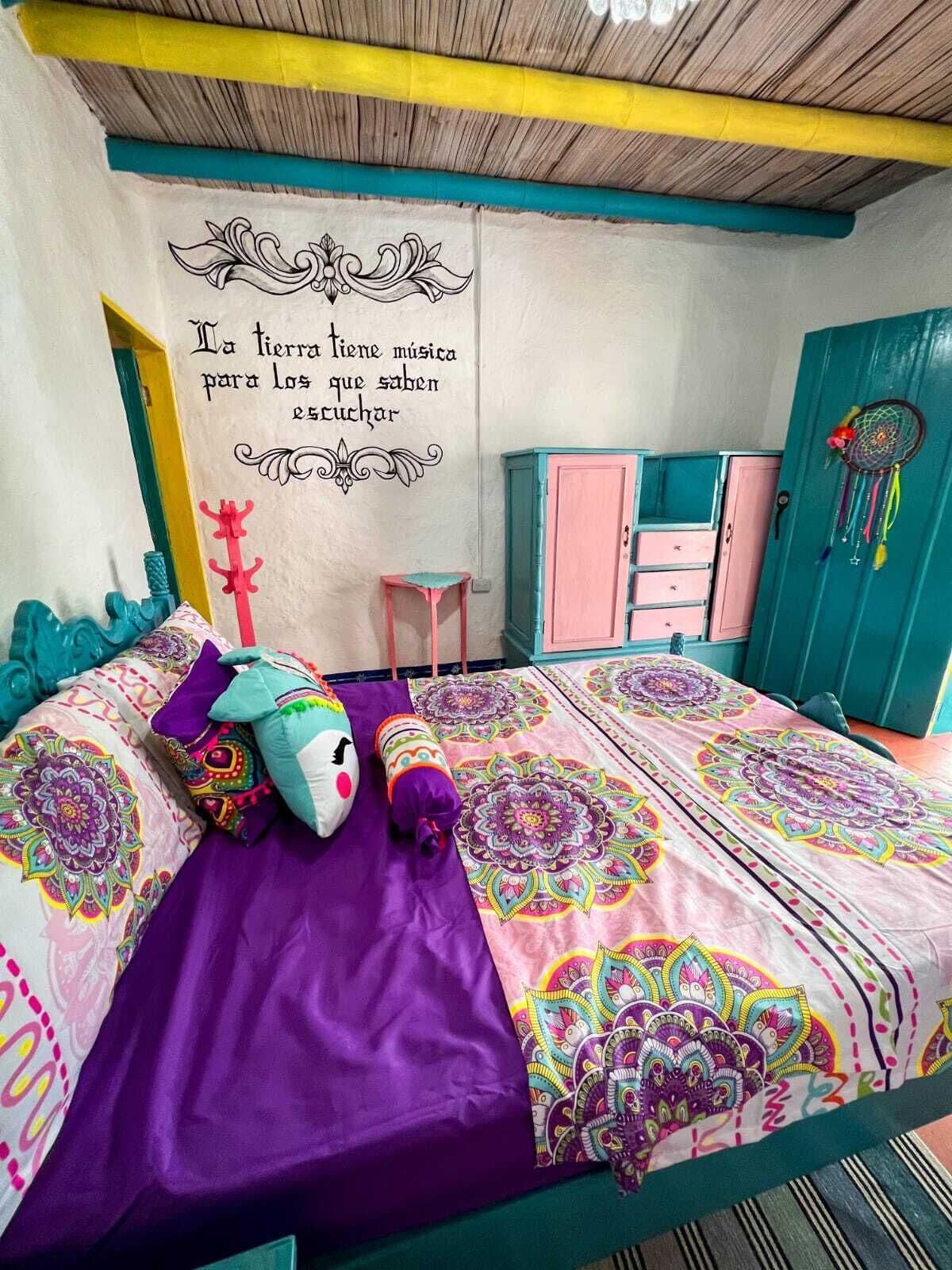 Une chambre à coucher avec un lit, une literie aux couleurs vives, un mur orné d’une citation décorative, un attrape-rêves et une petite table.