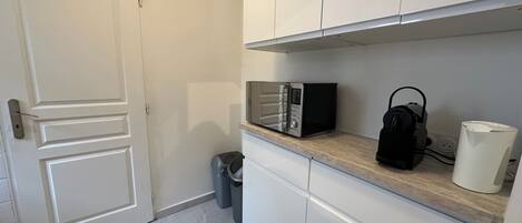 Una pequeña kitchenette con gabinetes blancos, microondas, cafetera y un cesto de basura.