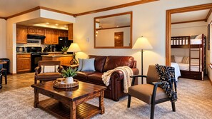 A living room with a brown leather sofa, a wooden coffee table, and a chair with a blanket. A kitchen is visible in the background with wooden cabinets and a microwave. - Snowed Inn Brian Head Condo Sleeps 6 (Brian Head)