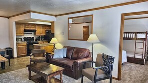 A living room with a brown leather sofa, a wooden coffee table, a patterned armchair, and a dining table. There is a kitchen visible in the background with wooden cabinets and a microwave. - Snowed Inn Brian Head Condo Sleeps 6 (Brian Head)