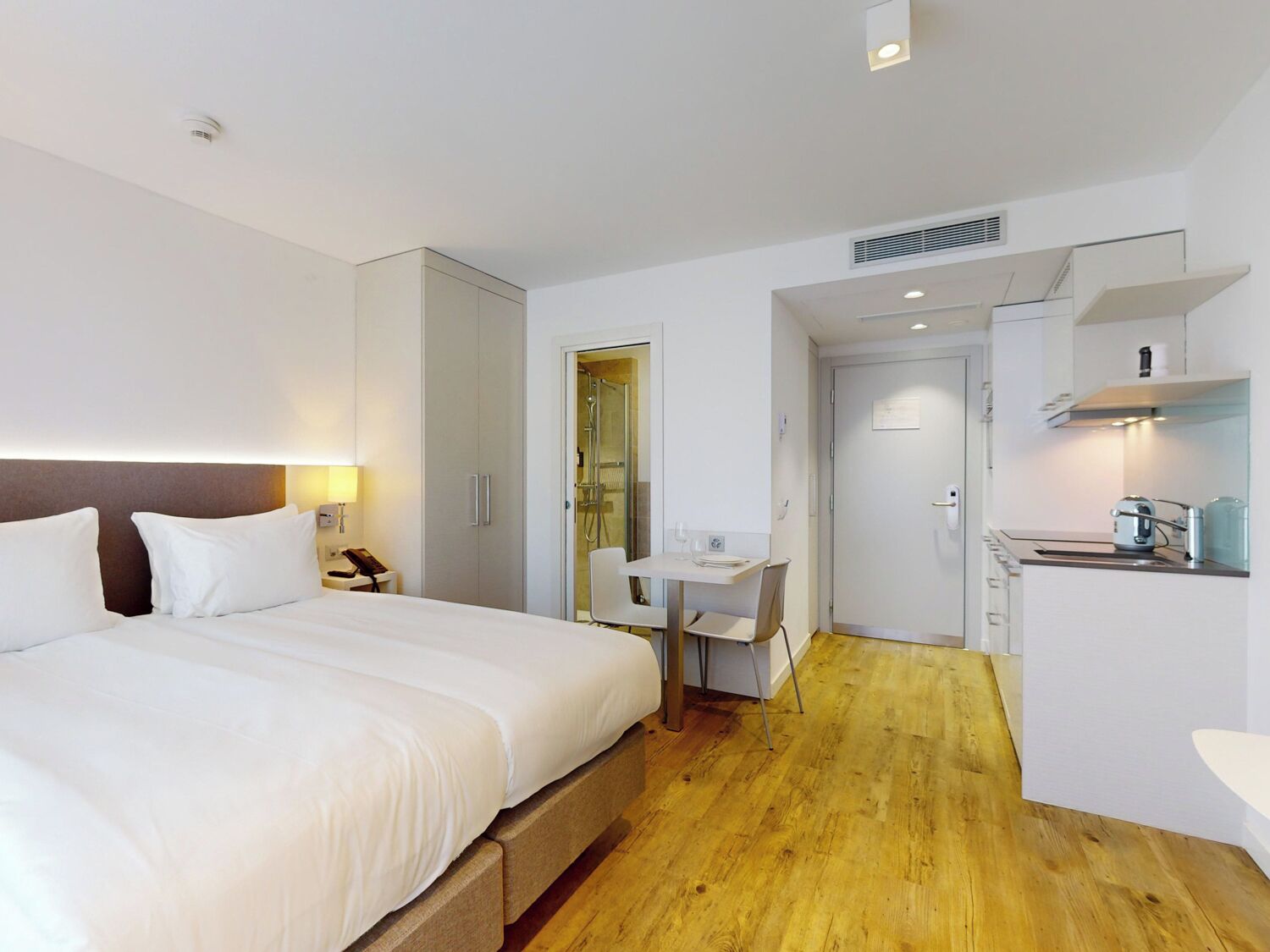 A hotel room with a large bed, a desk, a kitchenette, and a shower area.