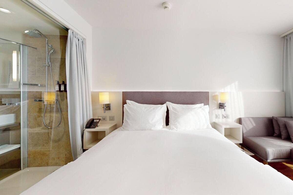 A hotel room with a large bed, a glass shower enclosure, a bedside table with a phone, and a seating area with a sofa.