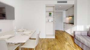 Una cocina moderna y minimalista con muebles blancos, una zona de comedor con mesa y sillas blancas, y un sofá gris en un espacio amplio y luminoso.