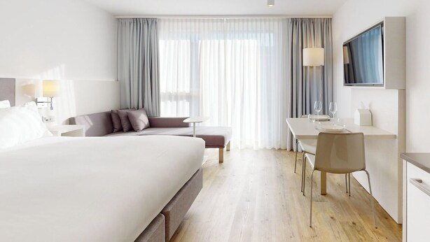 Una habitación de hotel moderna con una cama grande, un escritorio con silla, un televisor y una ventana con cortinas.