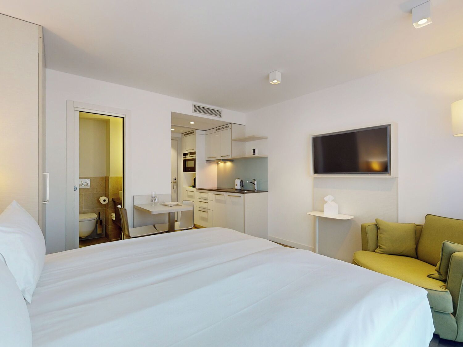 A hotel room with a large bed, a small kitchenette, a TV mounted on the wall, and a sofa.