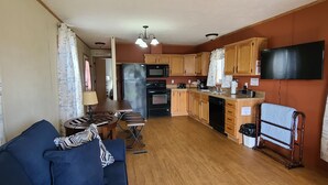 A compact kitchen with wooden cabinets, a refrigerator, stove, and a microwave. There is a dining table with chairs and a sofa in the living area.