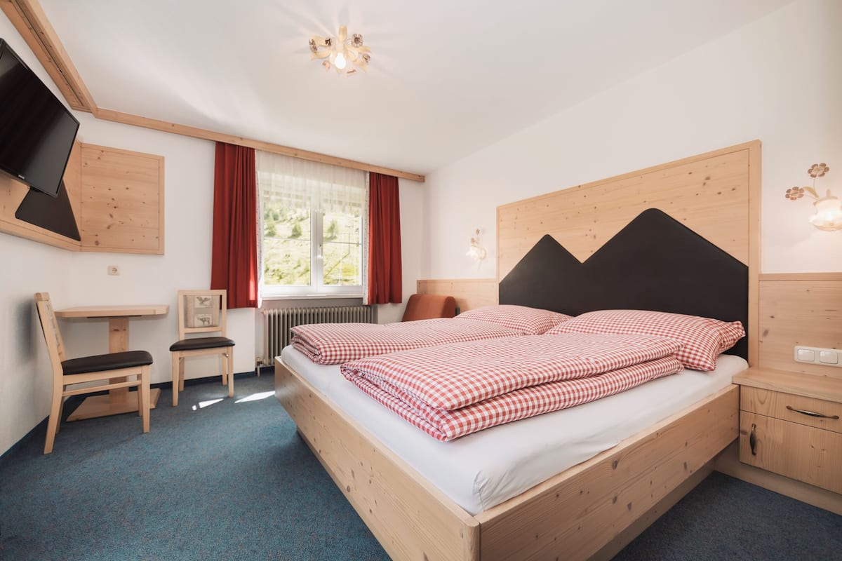 A bedroom with a wooden bed, a desk, a chair, a TV, and a window with curtains.