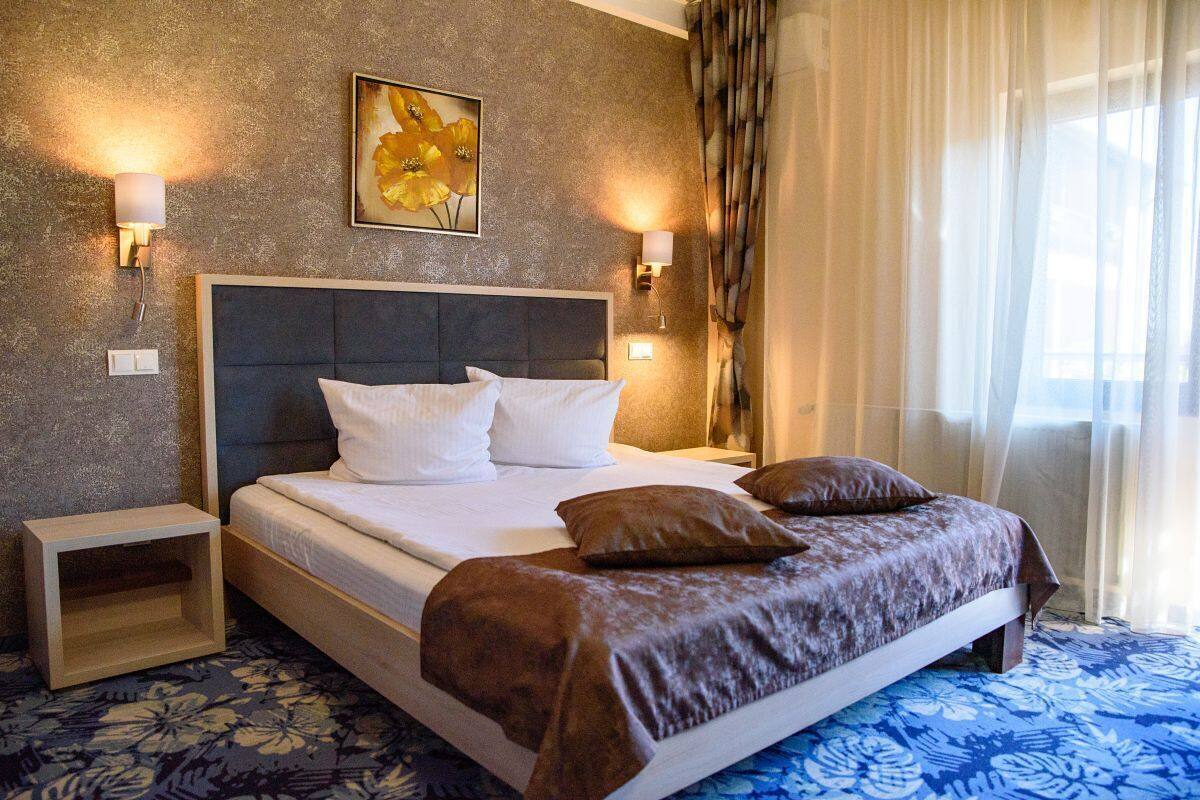 A hotel room with a large bed, two bedside tables, a wall-mounted lamp, and a window with curtains.