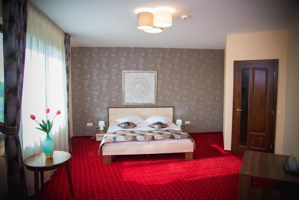 A hotel room with a bed, a nightstand, a vase with flowers, a red carpet, a wooden door, and a framed picture on the wall.