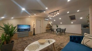 A modern living room with a blue sofa, a coffee table, a TV, and a dining area with chairs. - Hotel Stefani (Sibiu)