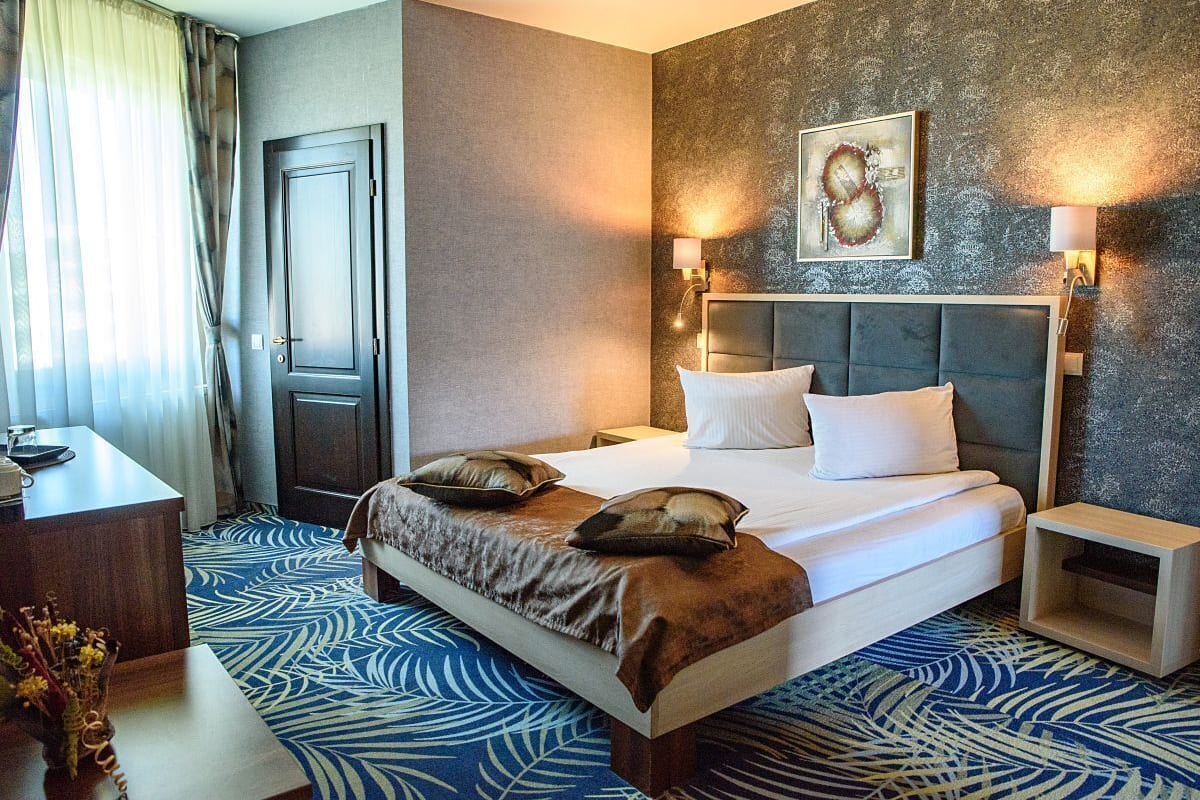 A hotel room with a large bed, two bedside tables, a desk, and a chair. There is a door and a window with curtains.