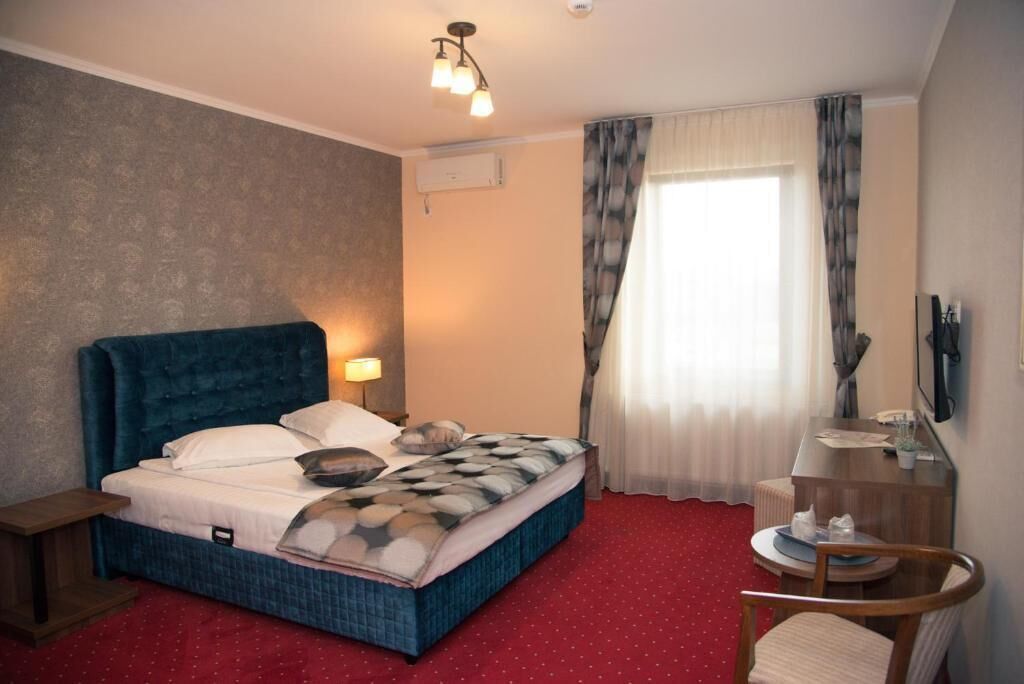 A hotel room with a large bed, a bedside table, a desk with a chair, a TV, and a window with curtains.