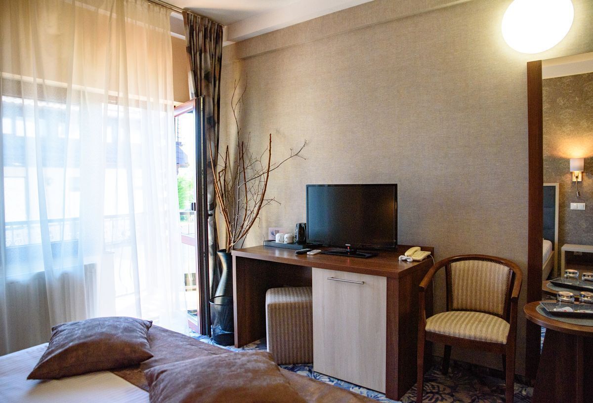 A hotel room with a bed, a desk with a television, a chair, and a small table with a lamp.