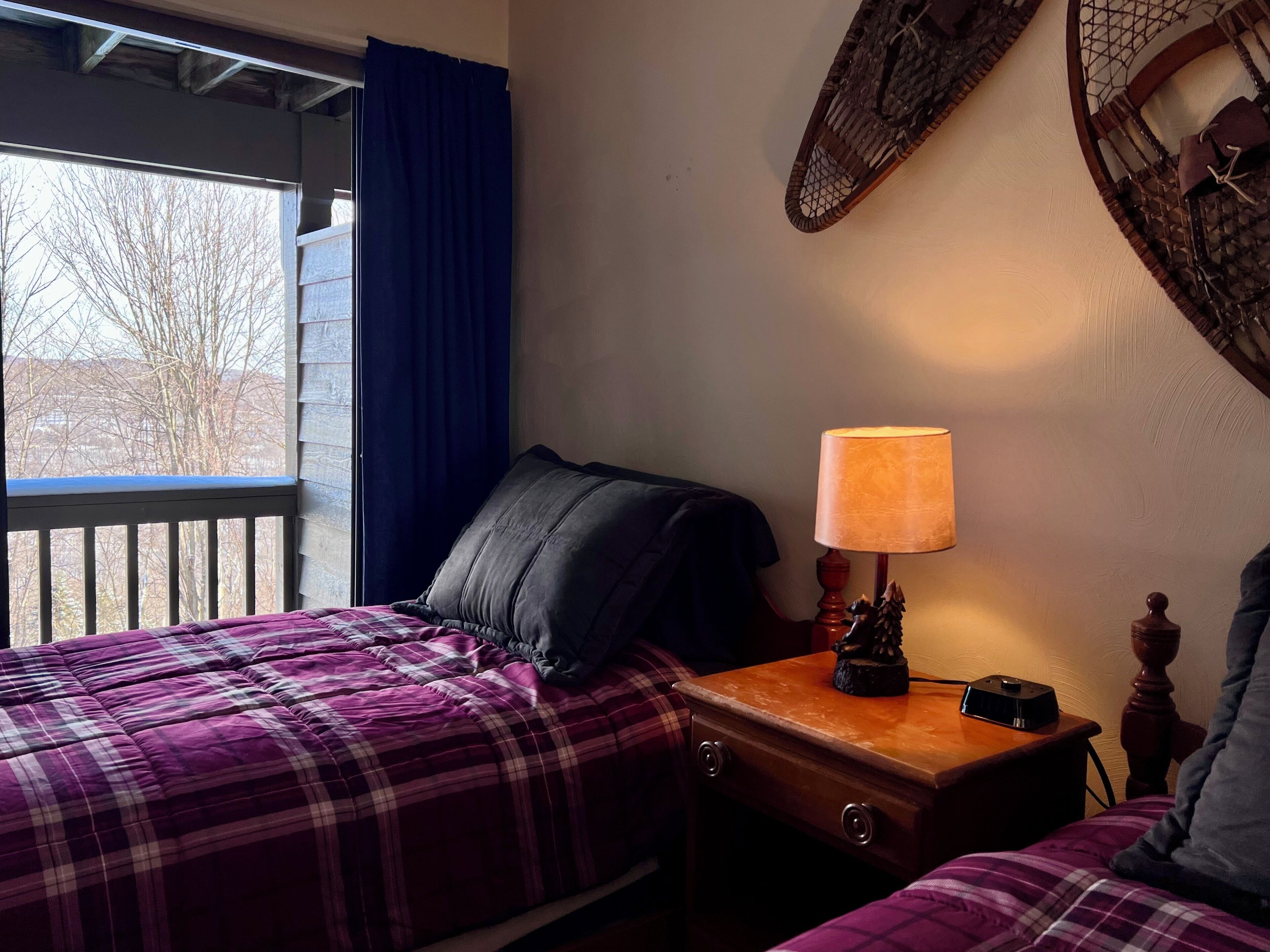 A bedroom with two beds, a nightstand with a lamp, and a view of trees through a window.