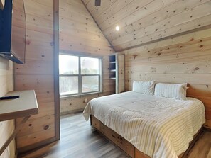 Une chambre avec un plafond en bois, un lit avec une literie à rayures, une fenêtre et un bureau avec une télécommande.