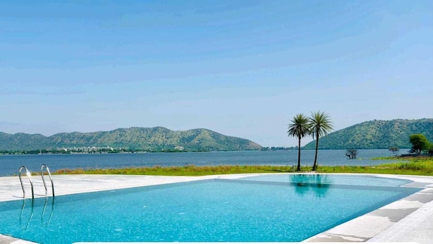A swimming pool with clear blue water, surrounded by a paved area and palm trees, with a view of mountains and a body of water in the background. - The Udaisarovar - Lakeside Paradise (Udaipur)