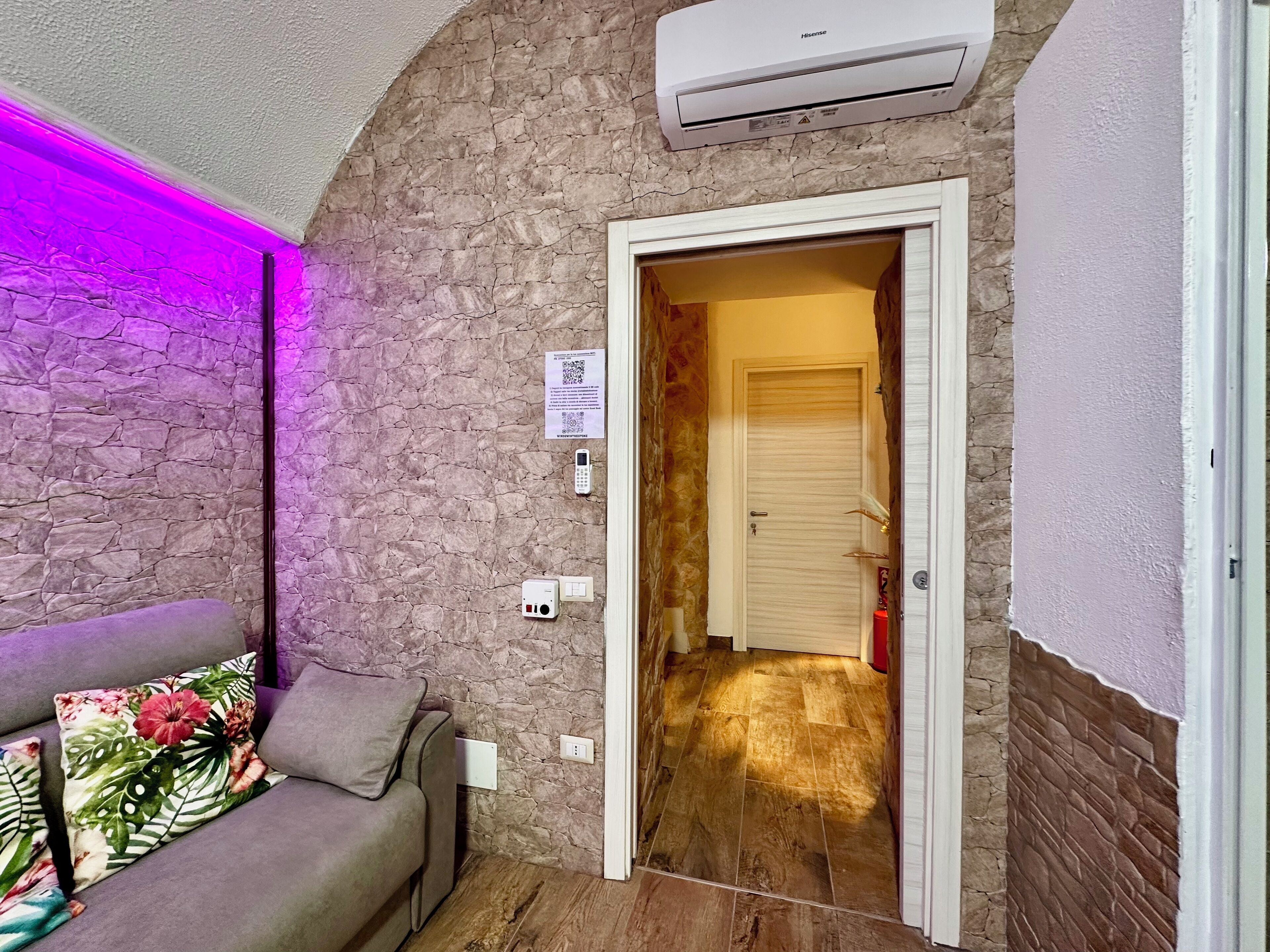 Um quarto com parede de pedra, um sofá cinza com almofadas florais, uma porta que dá para outro cômodo e um aparelho de ar condicionado fixado na parede.