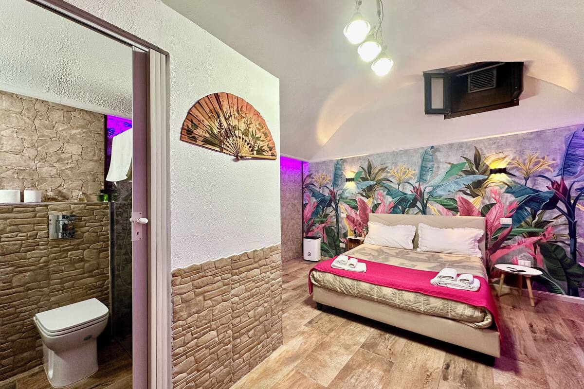 A bedroom with a bed, a wall-mounted fan, a bathroom with a toilet, and a shower area.