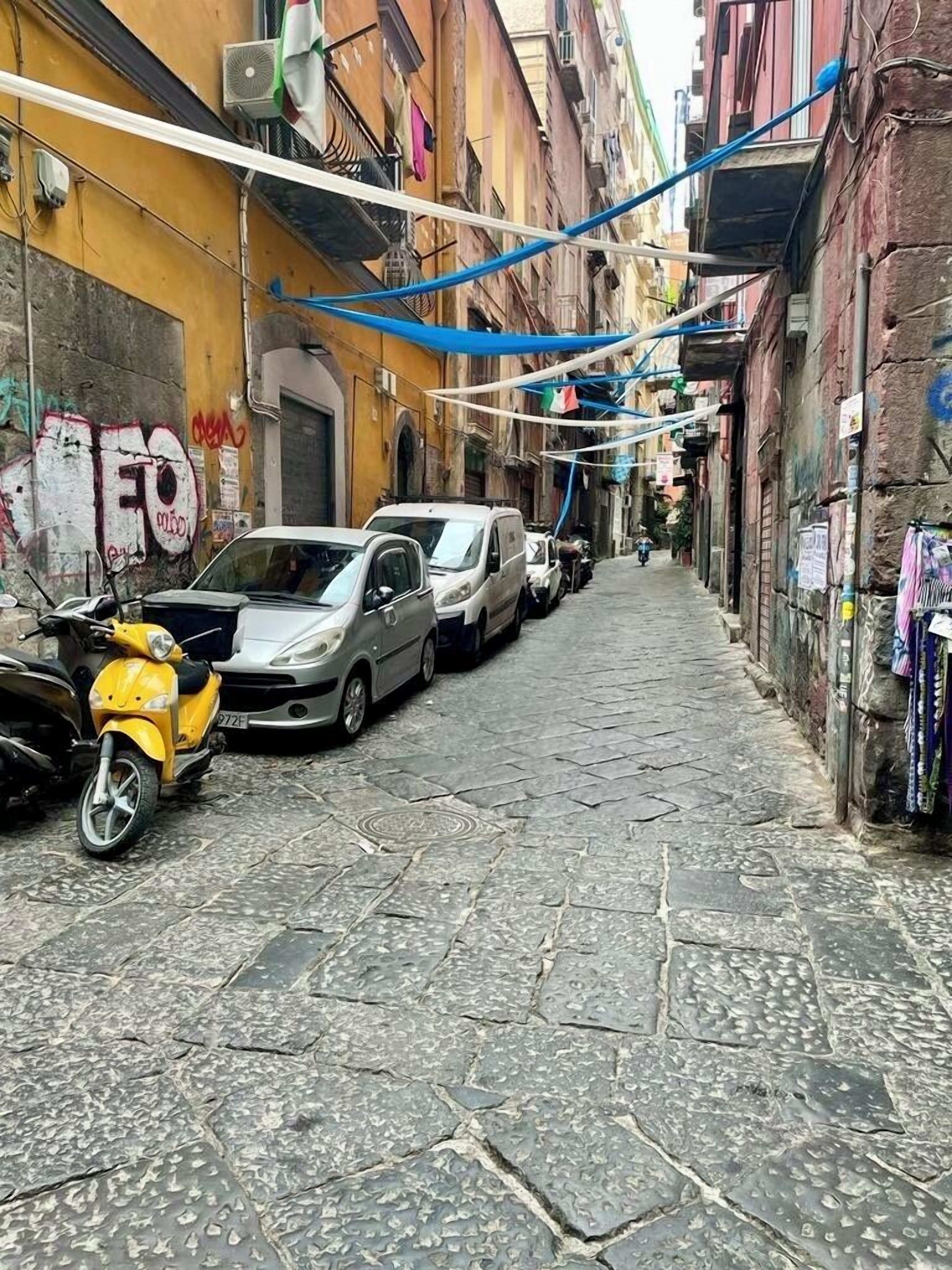 Une rue étroite bordée de voitures garées et d’un scooter, des immeubles dotés de balcons et une porte bien visible sur la gauche.