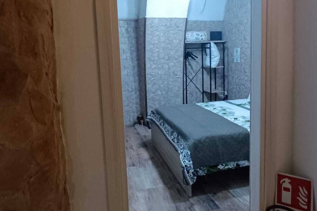 A bedroom with a bed, a small table, and a chair. There is a fire extinguisher on the wall.
