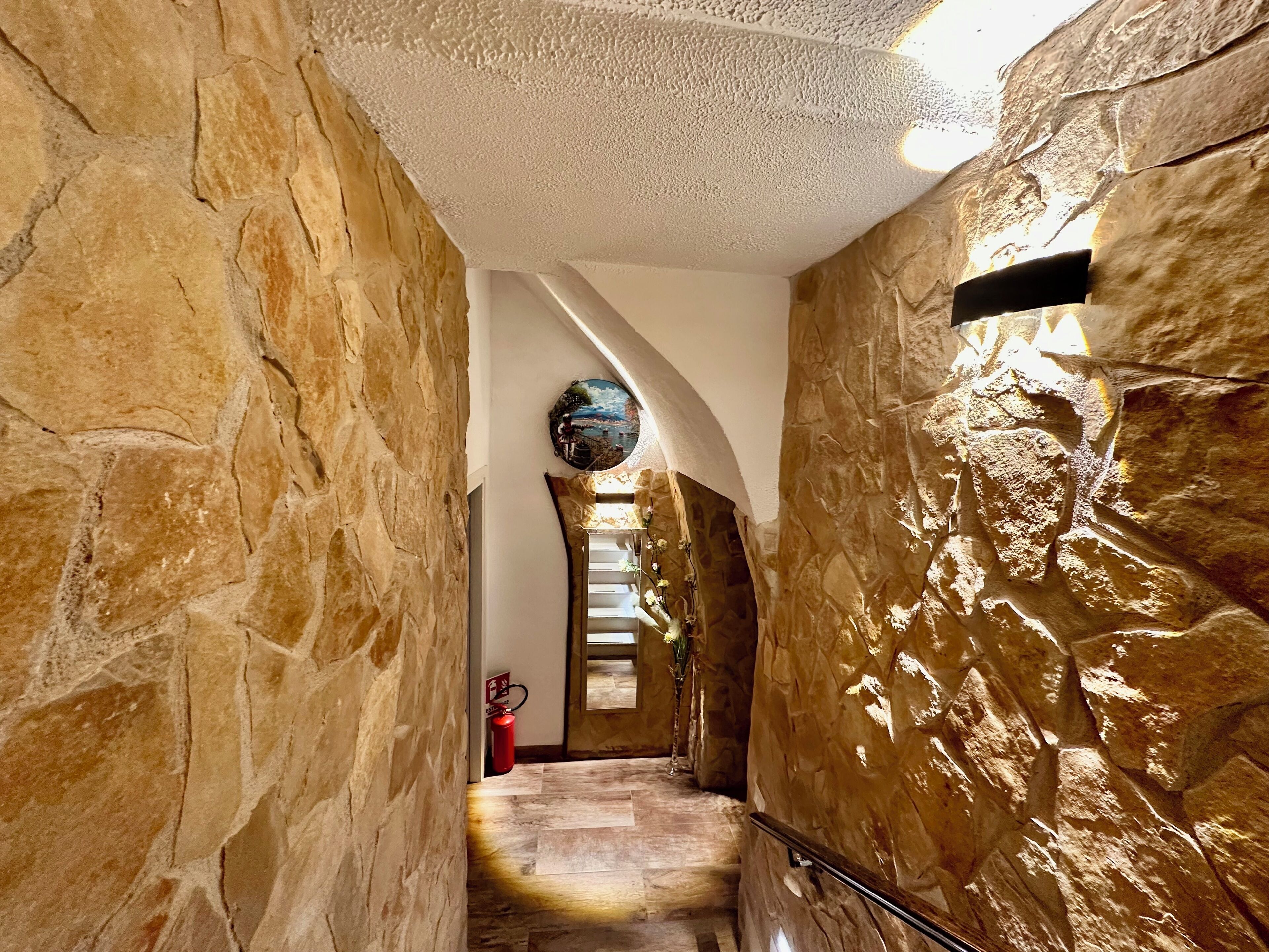 Un pasillo de paredes de piedra con un extintor, una obra de arte circular y una escalera que lleva a un nivel superior.