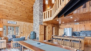 Interior rústico de cabana com uma mesa de jantar comprida, paredes de madeira, cozinha e um espaço de estar acolhedor.