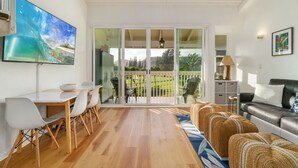 A spacious living area with a dining table, chairs, a sofa, and a large TV mounted on the wall. - Pu'uhonua Kai ~ Oceanside Refuge (Kahuku)