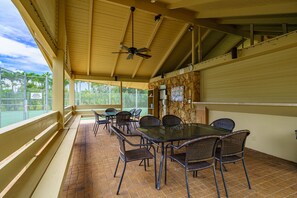 Een terras buiten met een tafel en stoelen, een plafondventilator en uitzicht op een tennisbaan.