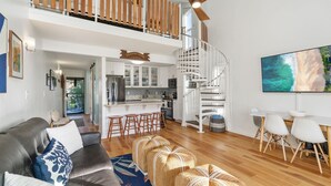 A spacious living area with a high ceiling, a kitchen with white cabinets, a dining area with a table and chairs, and a staircase leading to an upper level. - Pu'uhonua Kai ~ Oceanside Refuge (Kahuku)