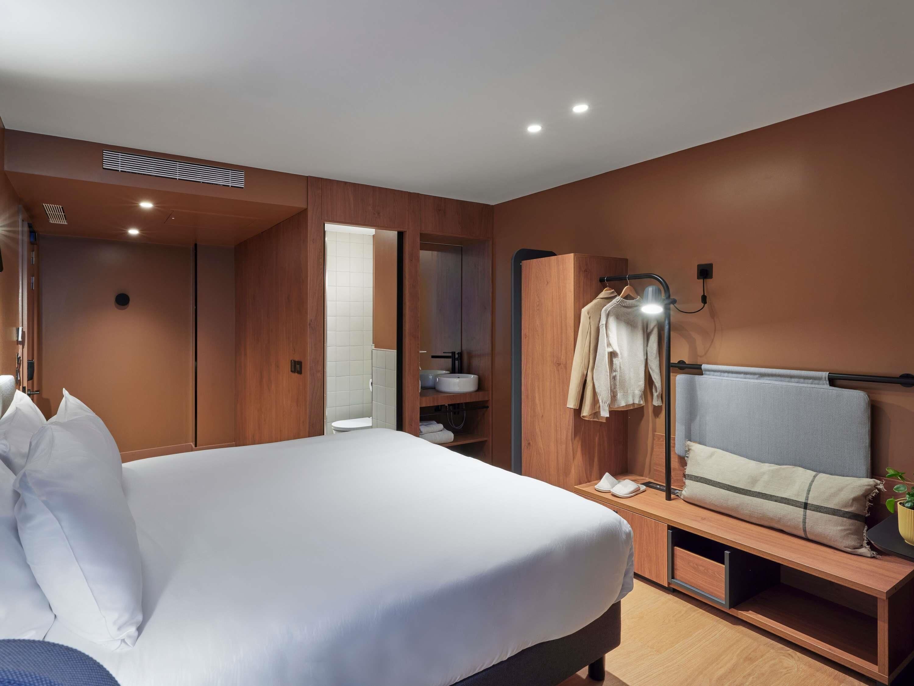 Une chambre d’hôtel moderne avec un grand lit, une armoire en bois, une table de chevet et un miroir.