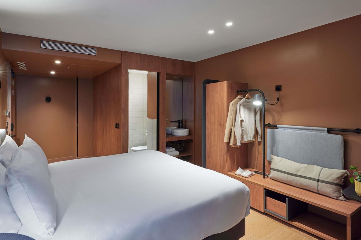 Moderni hotellihuone, jossa on suuri sänky, puinen vaatekaappi, yöpöytä ja peili.