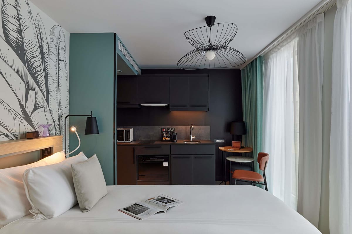 Et hotelværelse med seng, lille tekøkken, spiseplads og stort vindue med gardiner.
