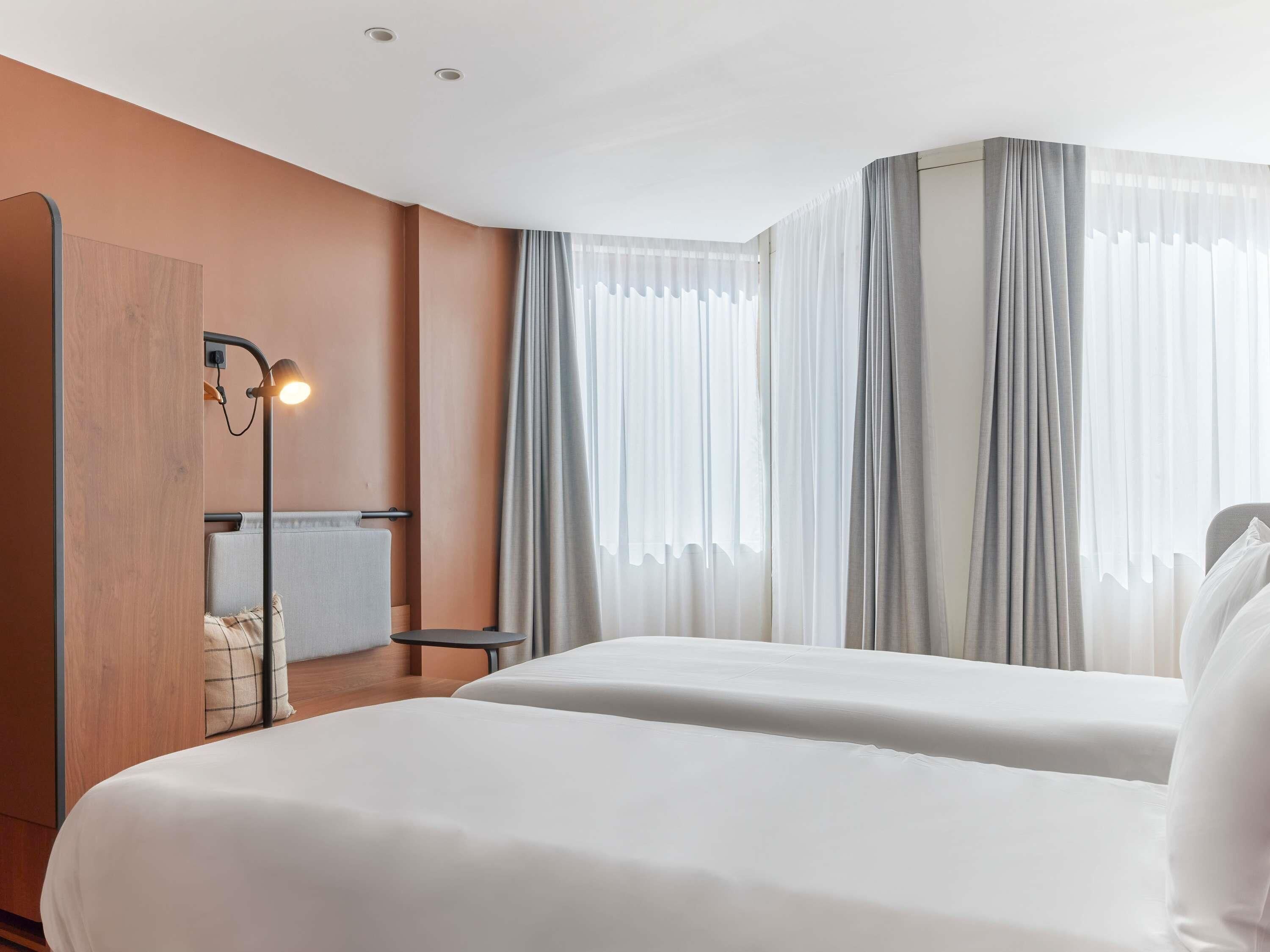 Et hotelværelse med to senge, et natbord, en lampe og et stort vindue med gardiner.