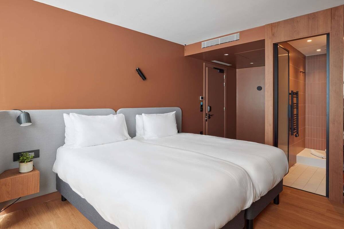 Una habitación de hotel moderna con una cama grande, una mesita de noche con lámpara, una planta pequeña y suelo de madera.