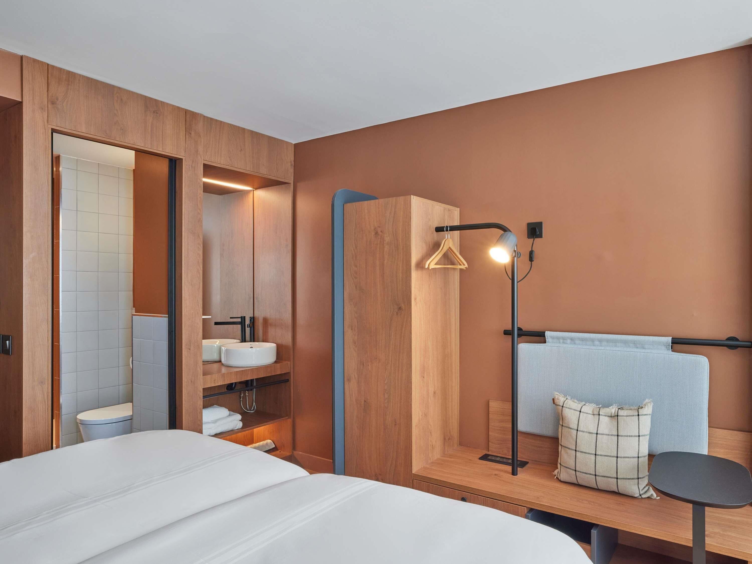 A modern hotel room with a large bed, a wooden wardrobe, a built-in sink, and a bedside table with a lamp.