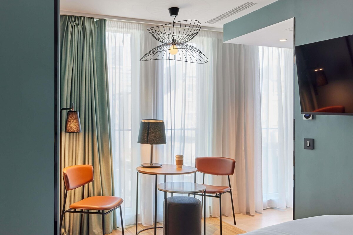 Hotellihuone, jossa on sänky, kaksi tuolia, pieni pöytä lampun kanssa, televisio ja ikkuna, jossa on verhot.