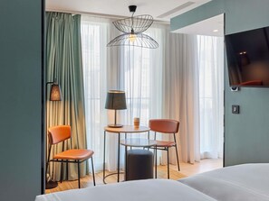 A hotel room with a bed, two chairs, a small table with a lamp, a TV, and a window with curtains. - Aparthotel Adagio London City East (London)