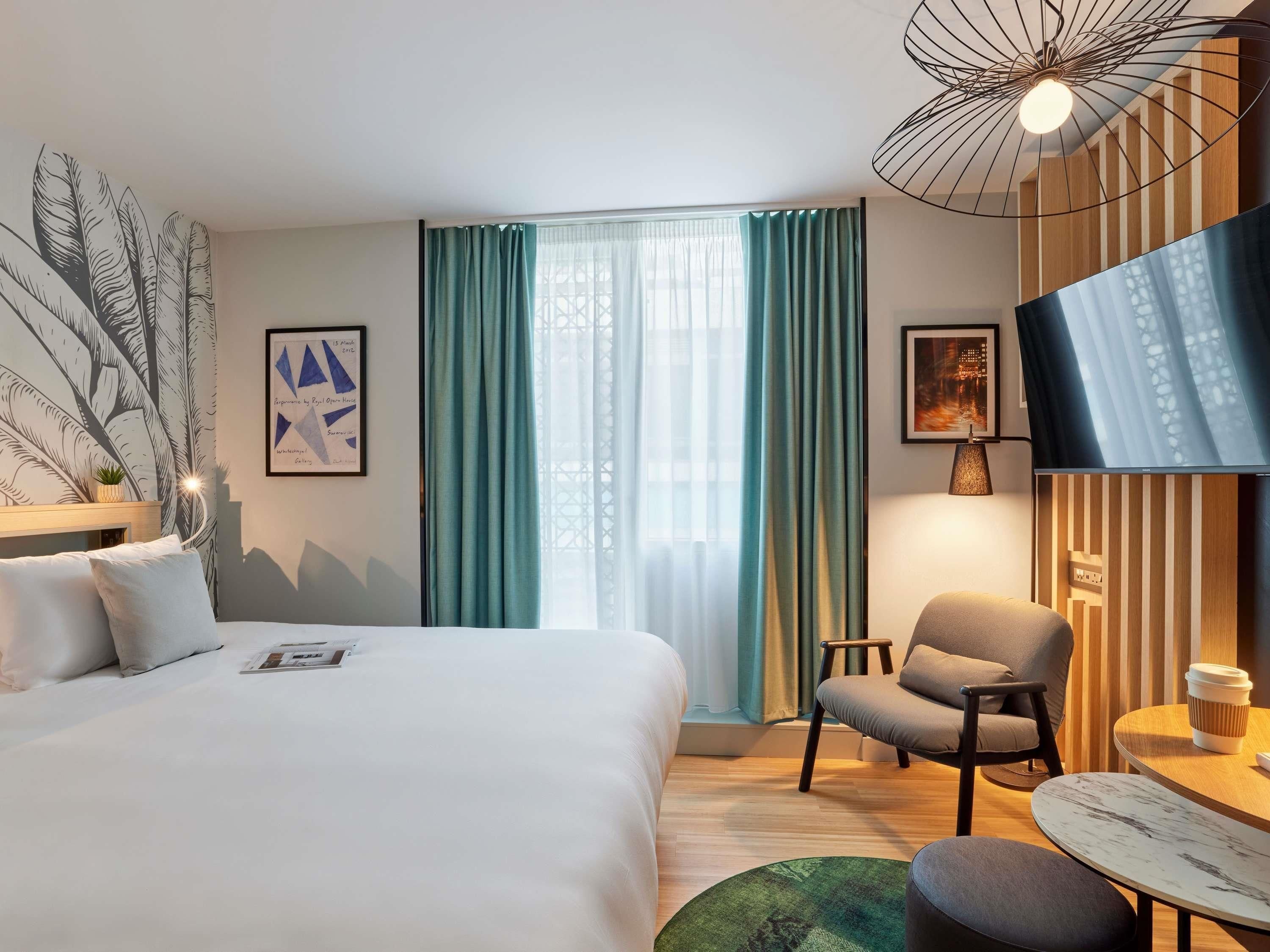 A hotel room with a bed, a chair, a table, and a large window with curtains.