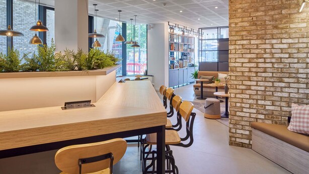 Een modern interieur met een lange houten tafel, zwarte stoelen, hangende pendantlampen, een accentmuur van baksteen, en een boekenplank vol boeken.