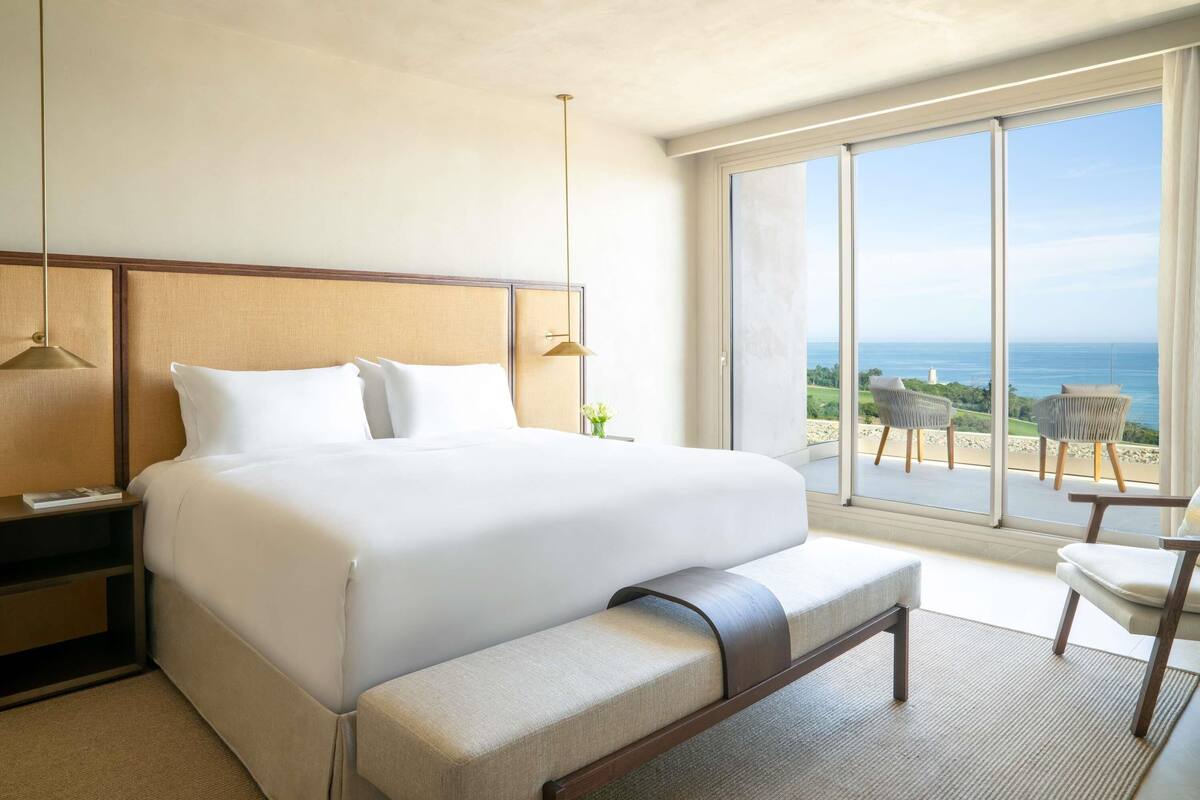 A hotel room with a large bed, a bench, a nightstand, a large window with a view of the sea, and a chair by the window.
