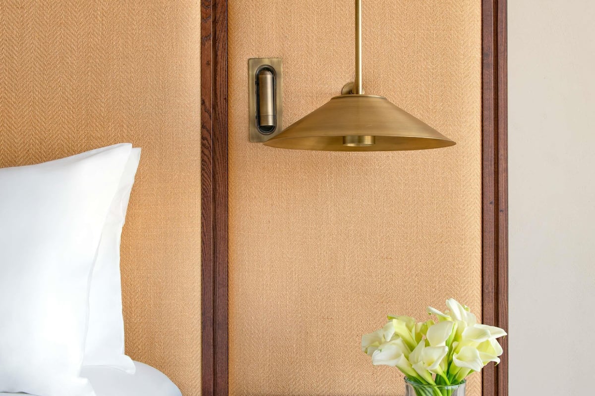 Habitación de hotel con pared beige texturizada, panel de madera, almohada blanca, lámpara colgante de latón y un jarrón con flores blancas.