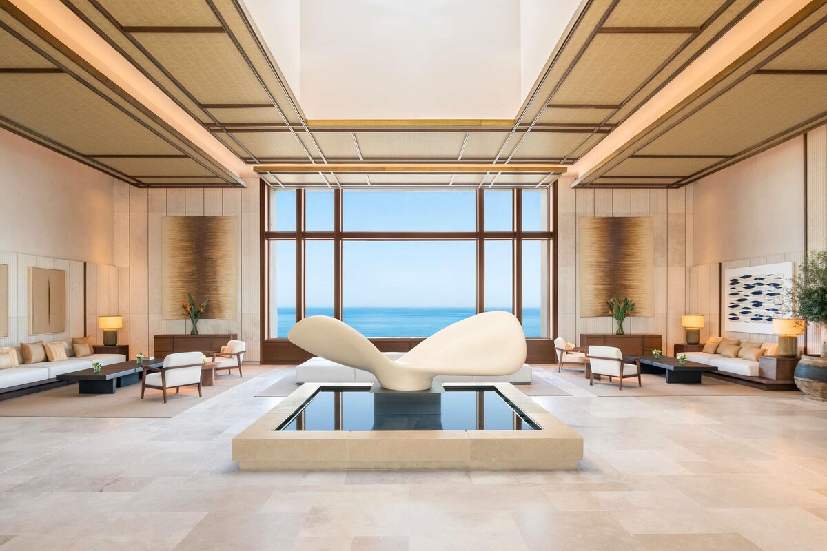 Eine großzügige Lobby mit einem großen Fenster mit Meerblick, einer zentralen Sitzecke und einem modernen Design mit Holzvertäfelung und hoher Decke.