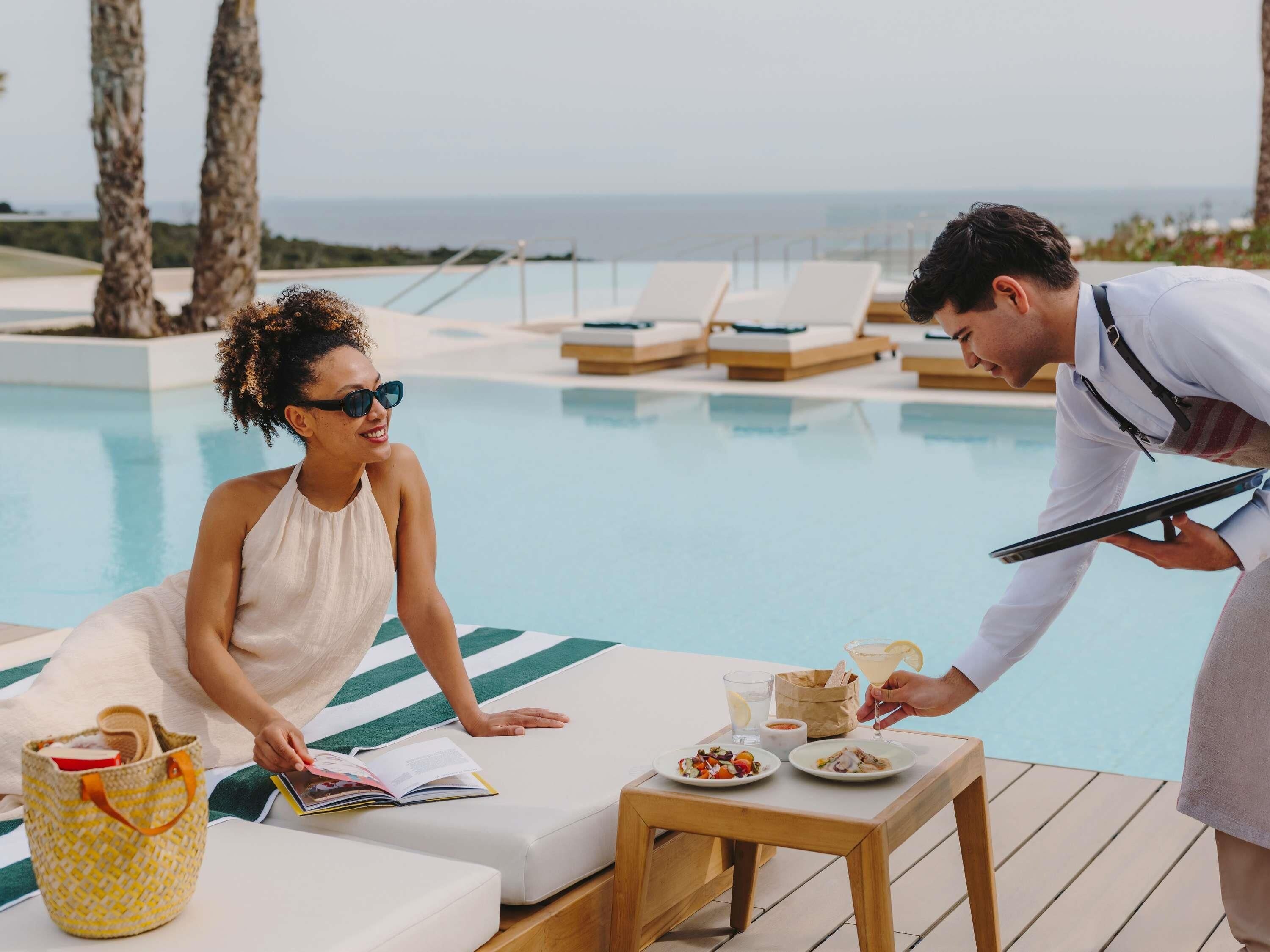 Escena junto a la piscina con sillones, una mesa con comida y bebidas, y una persona atendiendo.