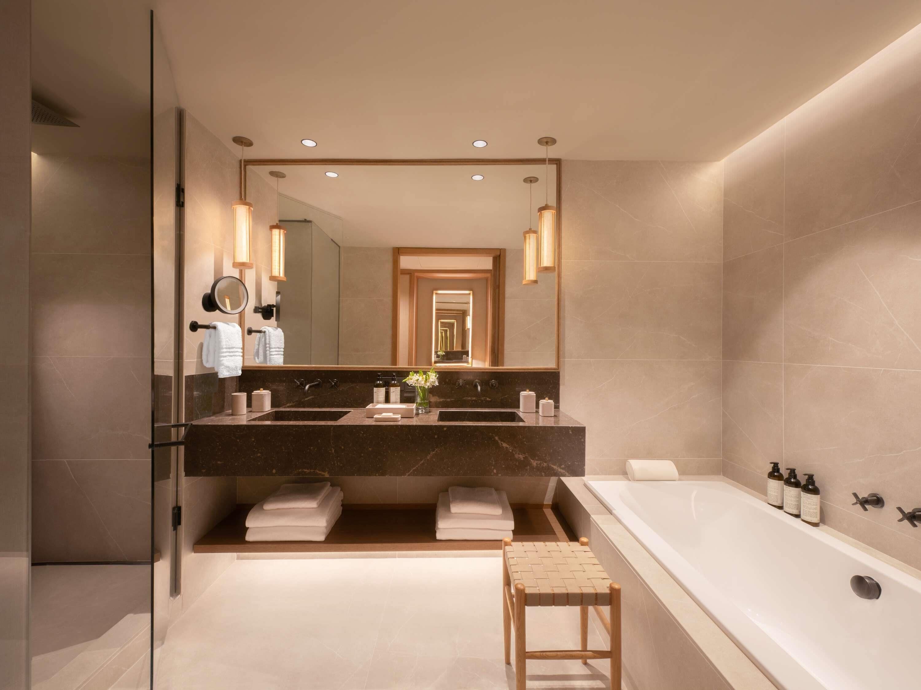 A modern bathroom with a large mirror, two sinks, a bathtub, and a small stool.
