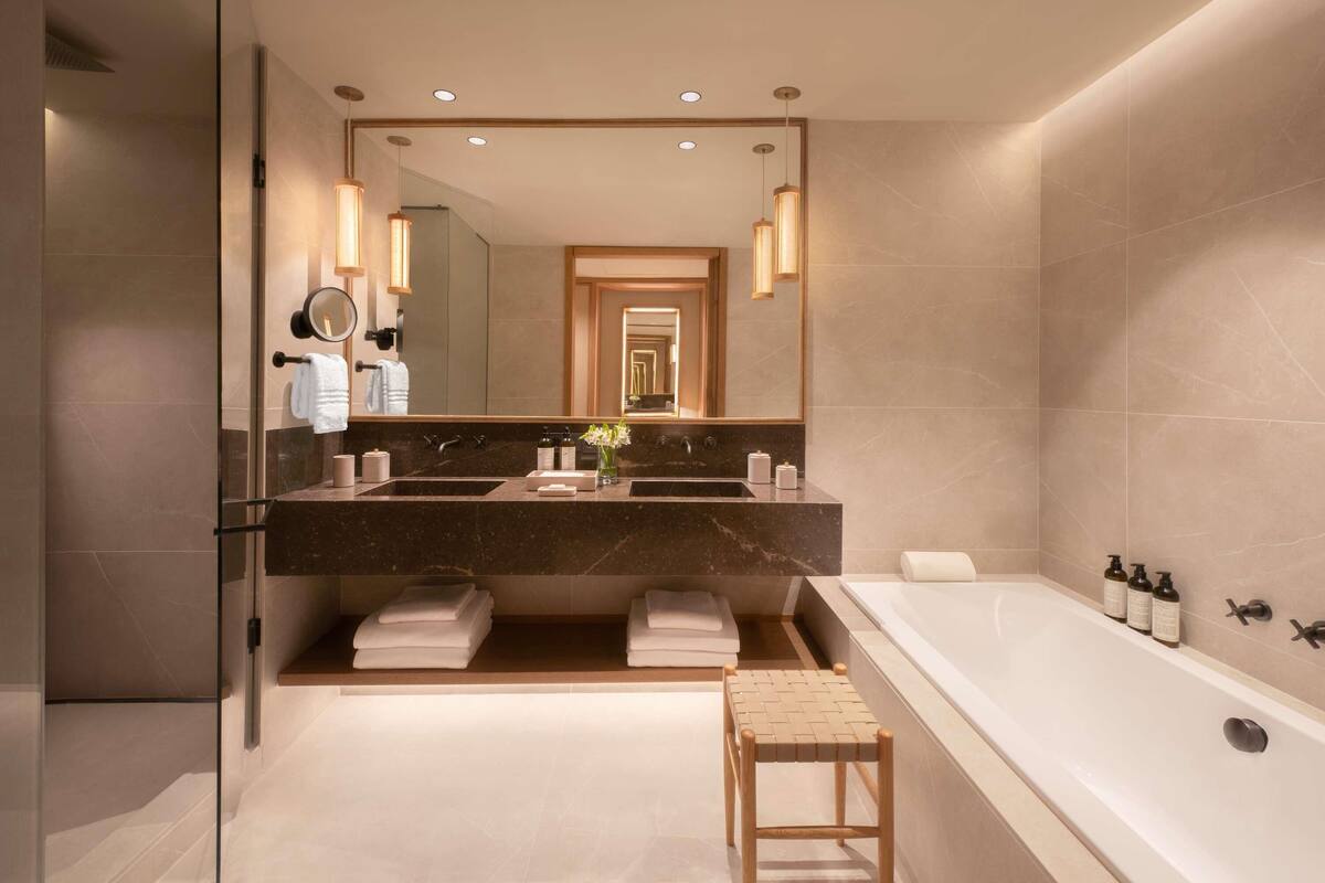A modern bathroom with a large mirror, two sinks, a bathtub, and a small stool.