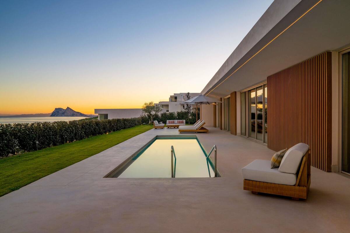 Un área de piscina moderna al aire libre con sillones de descanso y vistas al océano al atardecer.