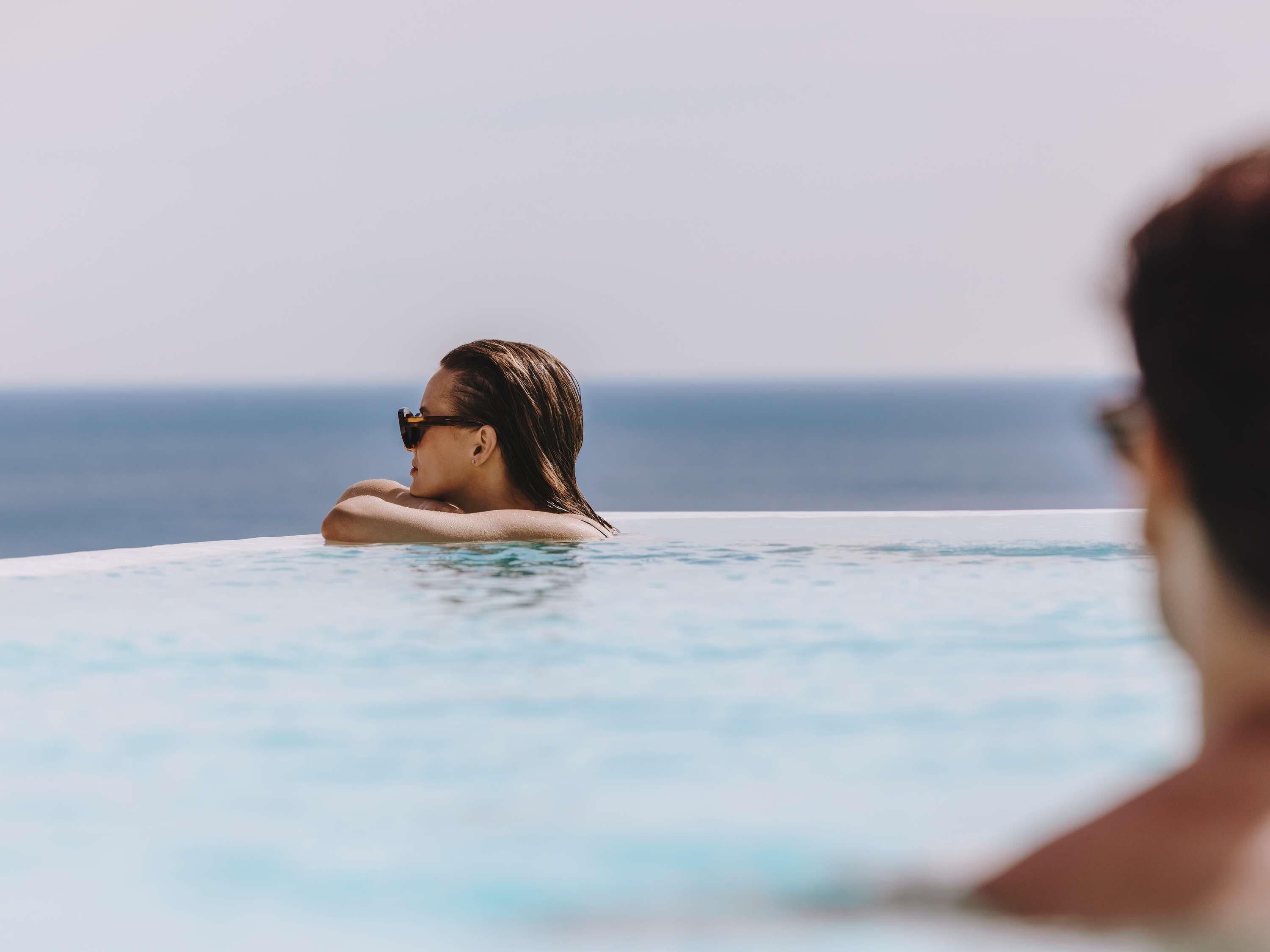 En person som kopplar av i en vacker, turkosblå pool som ser ut att sträcka sig oändligt långt, med utsikt över havet.