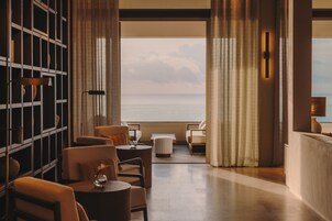 Un interior moderno con vistas al mar, que incluye una zona de estar con mesas y sillas redondas, una pequeña mesa redonda con un jarrón y una estantería.